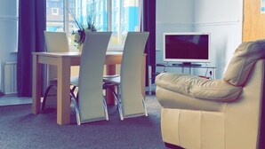 A modern room with a dining table, chairs, a sofa, a TV, and a window with curtains. - Henson Hotel (Blackpool)