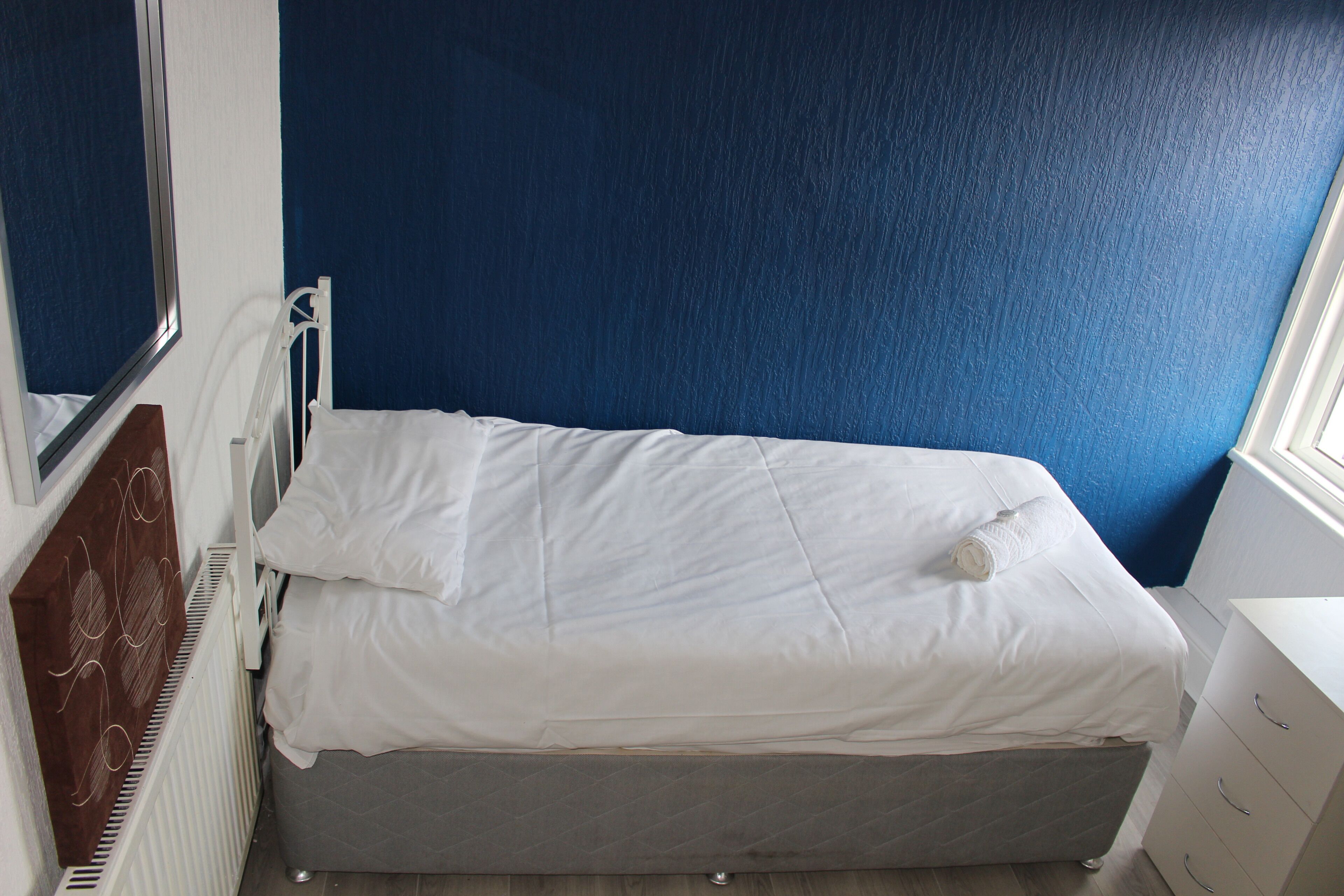 Un petit lit bien fait, avec une literie blanche, dans une pièce aux murs bleus et dotée d’une fenêtre.