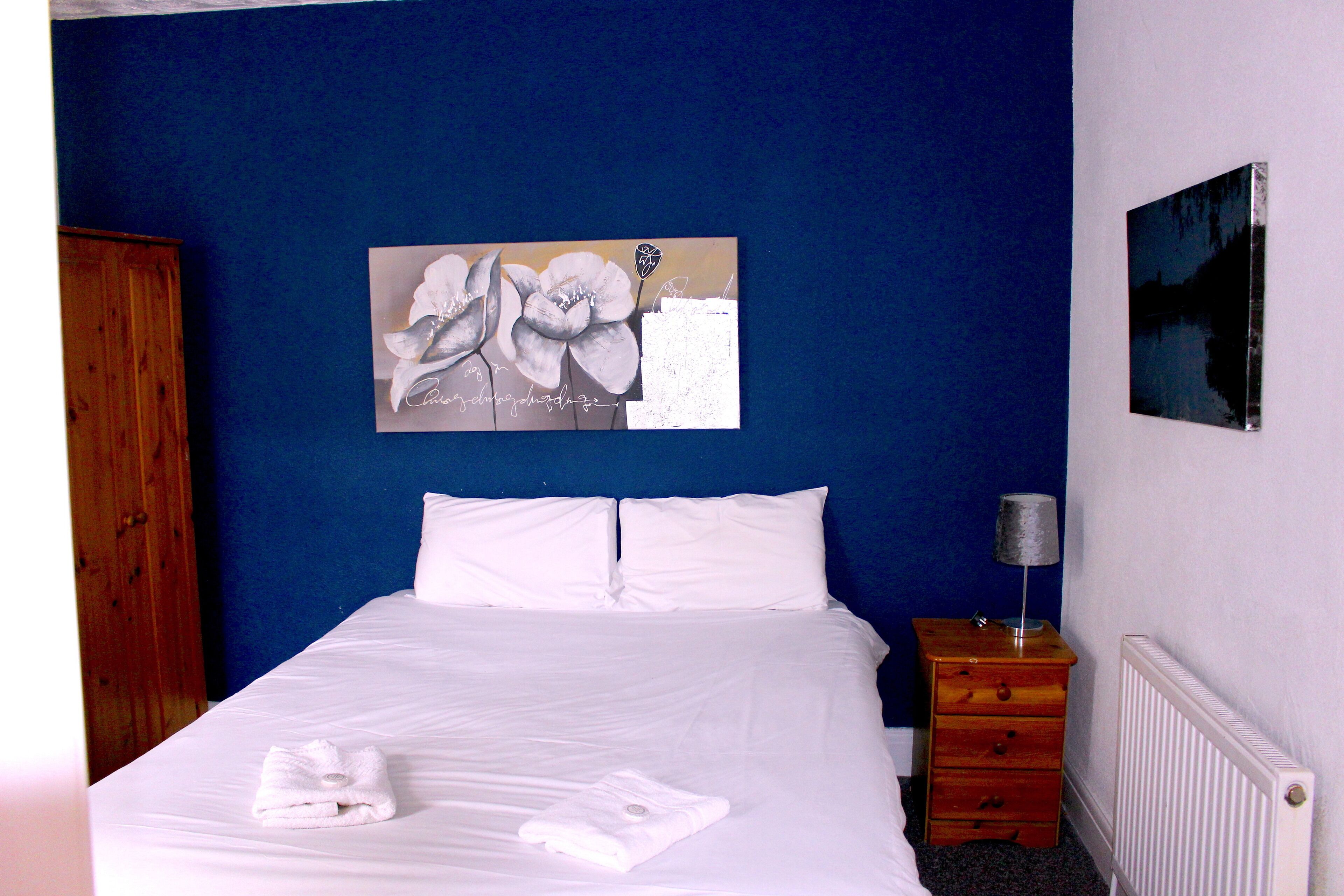 A neatly made bed with white linens and towels, a wooden nightstand with a lamp, and a painting on the wall.