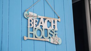Une enseigne en bois portant l’inscription « Bienvenue à la maison de plage » est accrochée à un mur bleu.
