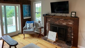 Ein gemütliches Wohnzimmer mit Kamin, einem darüber montierten Fernseher und einem Sessel mit Kopfkissen im Cape-Cod-Stil.