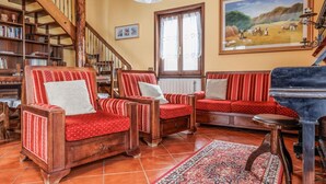 A cozy living room with two red upholstered chairs, a wooden piano, and a bookshelf filled with books. - Appartamento Panoramico Edolo (Vico)