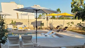 An outdoor grill area with skewers of meat, surrounded by lounge chairs and umbrellas, with a pool and palm trees in the background. -  Beautiful villa with pool in Specchia (Specchia)