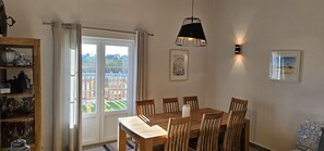 En matplats med ett träbord och stolar, en svart hänglampa och utsikt genom en glasdörr till en trädgård utomhus.