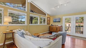 Un salón espacioso con un sofá grande, una mesa de centro de madera y un televisor. Varias ventanas ofrecen luz natural y vistas al exterior.