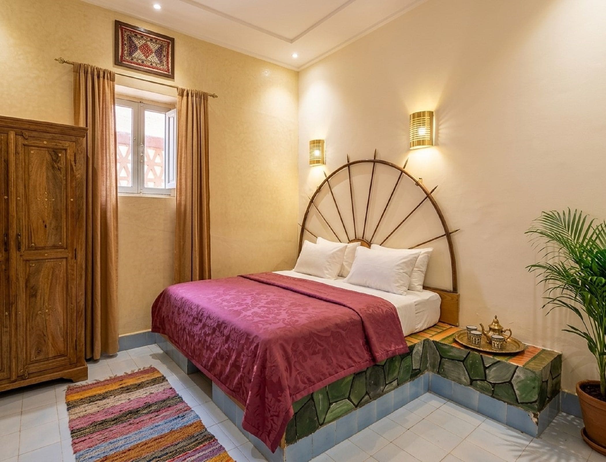 A bedroom with a large bed, a wooden wardrobe, a window with curtains, a wall-mounted light fixture, and a potted plant.