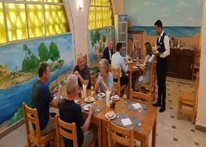A restaurant with wooden tables and chairs, a mural of a landscape, and a waiter serving guests. - Hllol Hotel Abu Simbel (Abu Simbel)