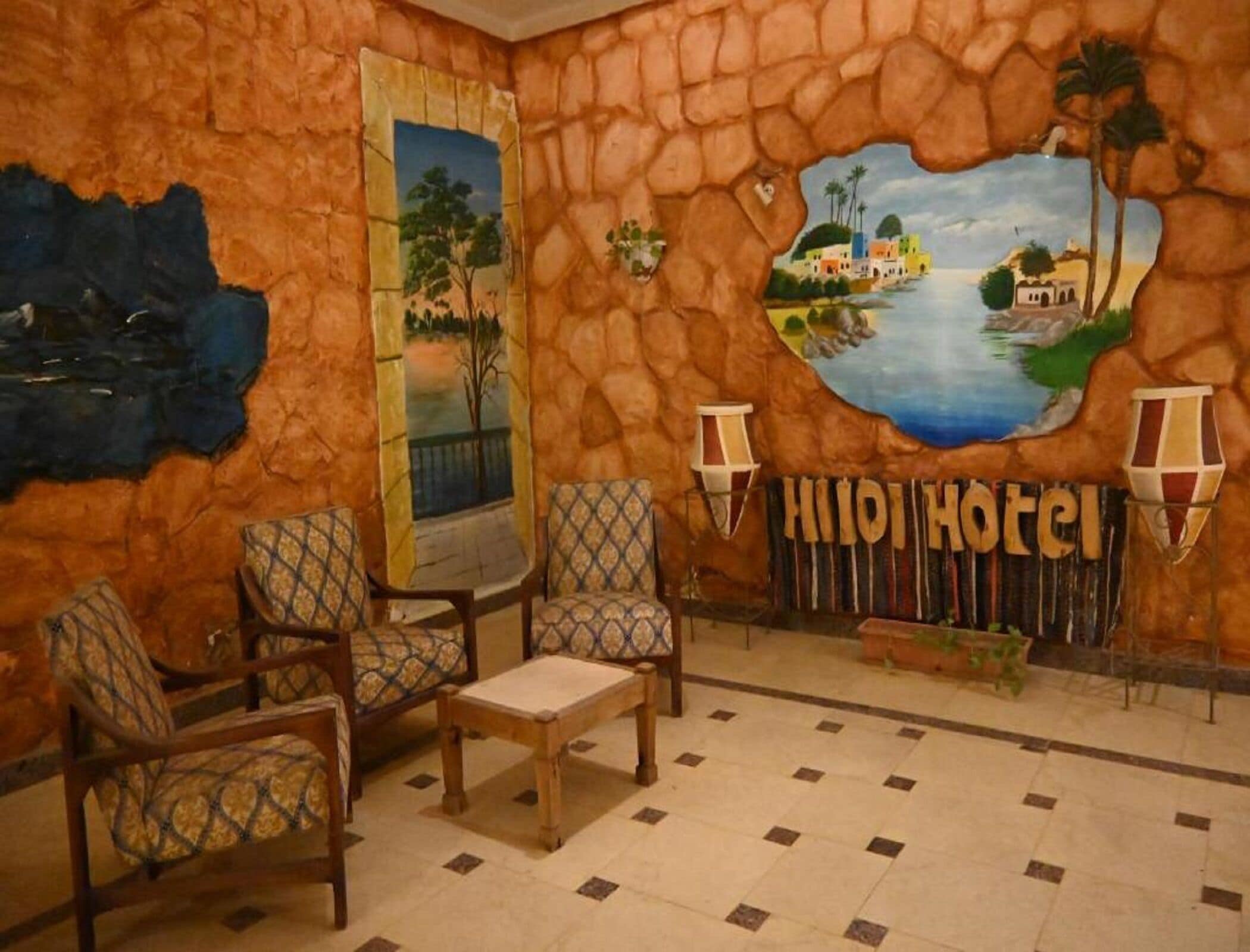 Eine Lobby mit Steinwänden, einem Gemälde einer Küstenszene, zwei Sesseln, einem kleinen Tisch und einem Schild mit der Aufschrift 'HIJHI Hotel'.