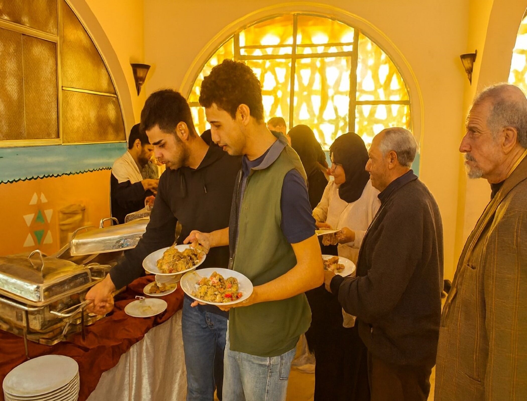 Eine Gruppe von Personen schöpft sich Essen an einem Buffet, im Hintergrund ist ein dekoratives Fenster zu sehen.