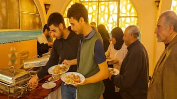 Eine Gruppe von Personen schöpft sich Essen an einem Buffet, im Hintergrund ist ein dekoratives Fenster zu sehen.