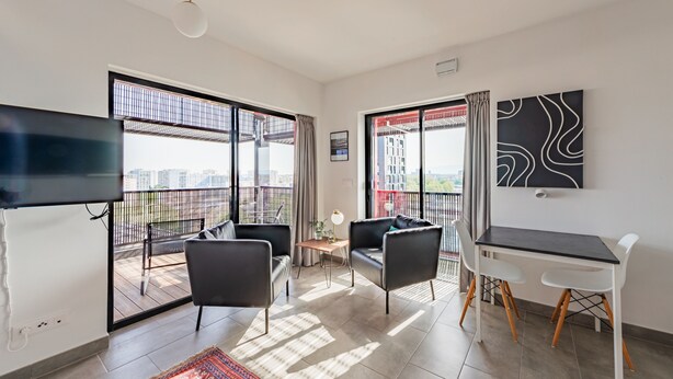 Ein modernes Wohnzimmer mit einem Flachbildfernseher, zwei schwarzen Sofas, einem kleinen Tisch und einem weißen Schreibtisch mit zwei Stühlen. Große Fenster bieten Blick auf die Stadt.