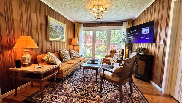 Un salon chaleureux avec un mur lambrissé en bois, une grande fenêtre, un téléviseur et un tapis à motifs.