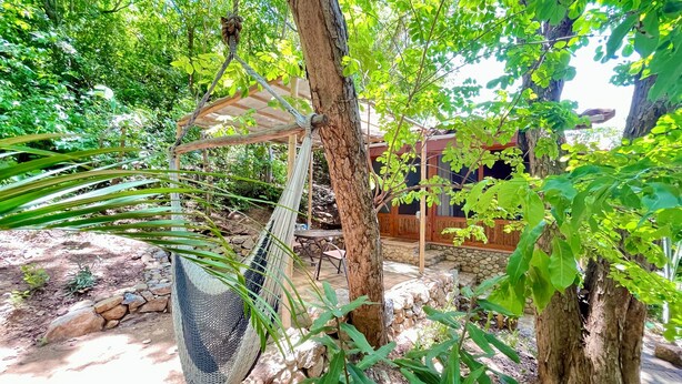 Una hamaca colgada de un árbol, una terraza de madera y un muro de piedra en un exuberante jardín.