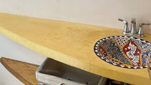Un baño con una ventana de listones de madera, un espejo montado en la pared, un lavabo con un salpicadero de azulejos decorativos y una pequeña cesta de mimbre.