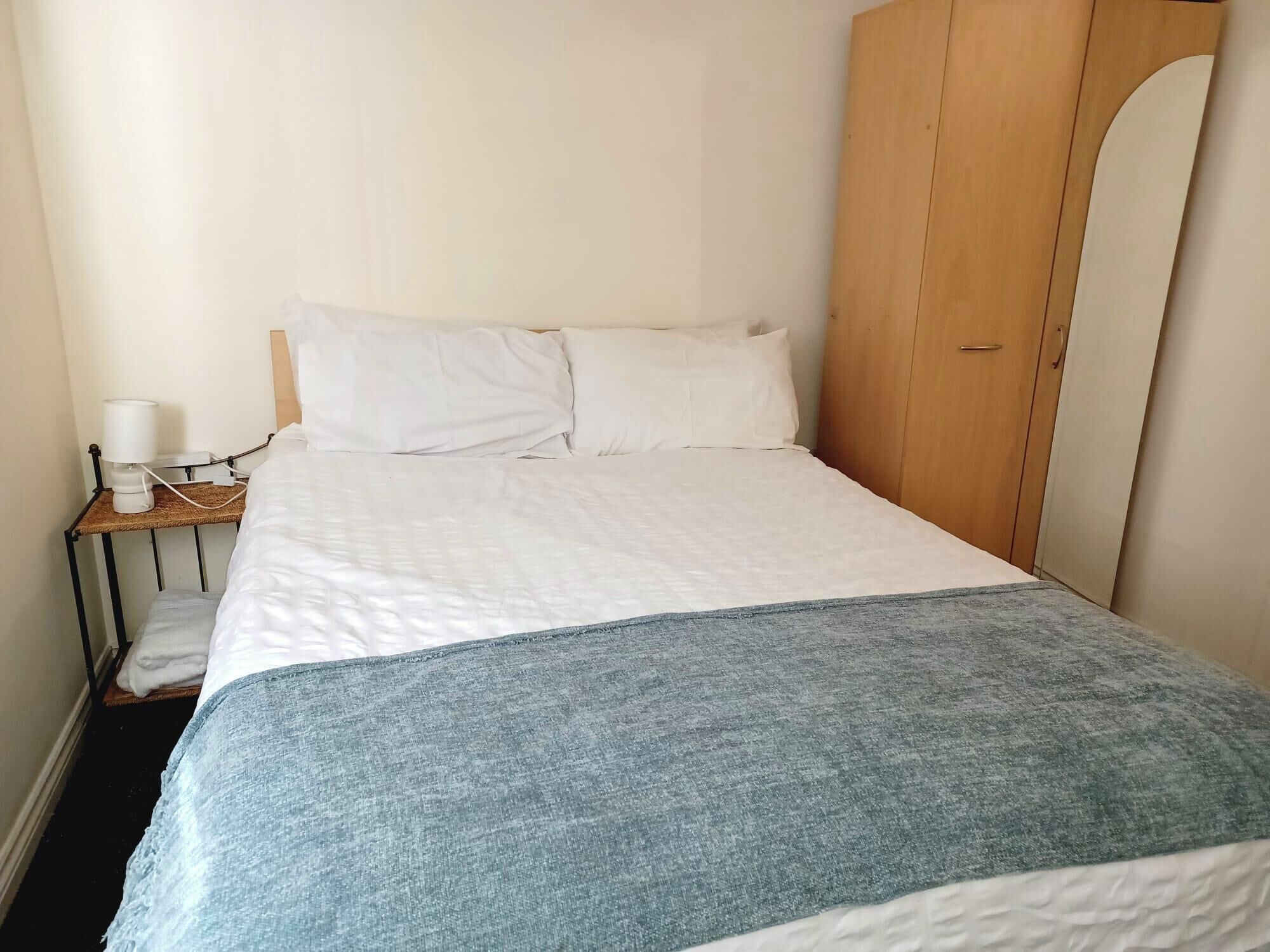 A single bed with a white duvet and a blue blanket, a bedside table with a lamp, and a wooden wardrobe.