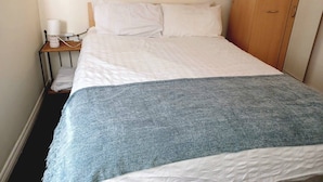 A single bed with a white and blue blanket, a bedside table with a lamp, and a wooden wardrobe.