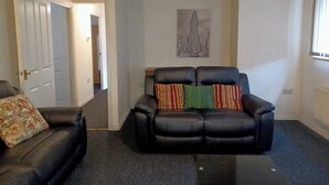 A living room with a black leather sofa, a floral-patterned armchair, and a black ottoman. There is a door leading to another room, a framed picture on the wall, and a window with blinds.