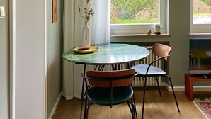Uma sala de jantar com uma mesa redonda, duas cadeiras, um vaso com flores secas e uma janela com vista para vegetação.