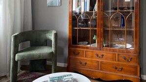 Quarto com um armário de madeira, uma cadeira verde, uma mesa redonda com revistas e um tapete estampado.