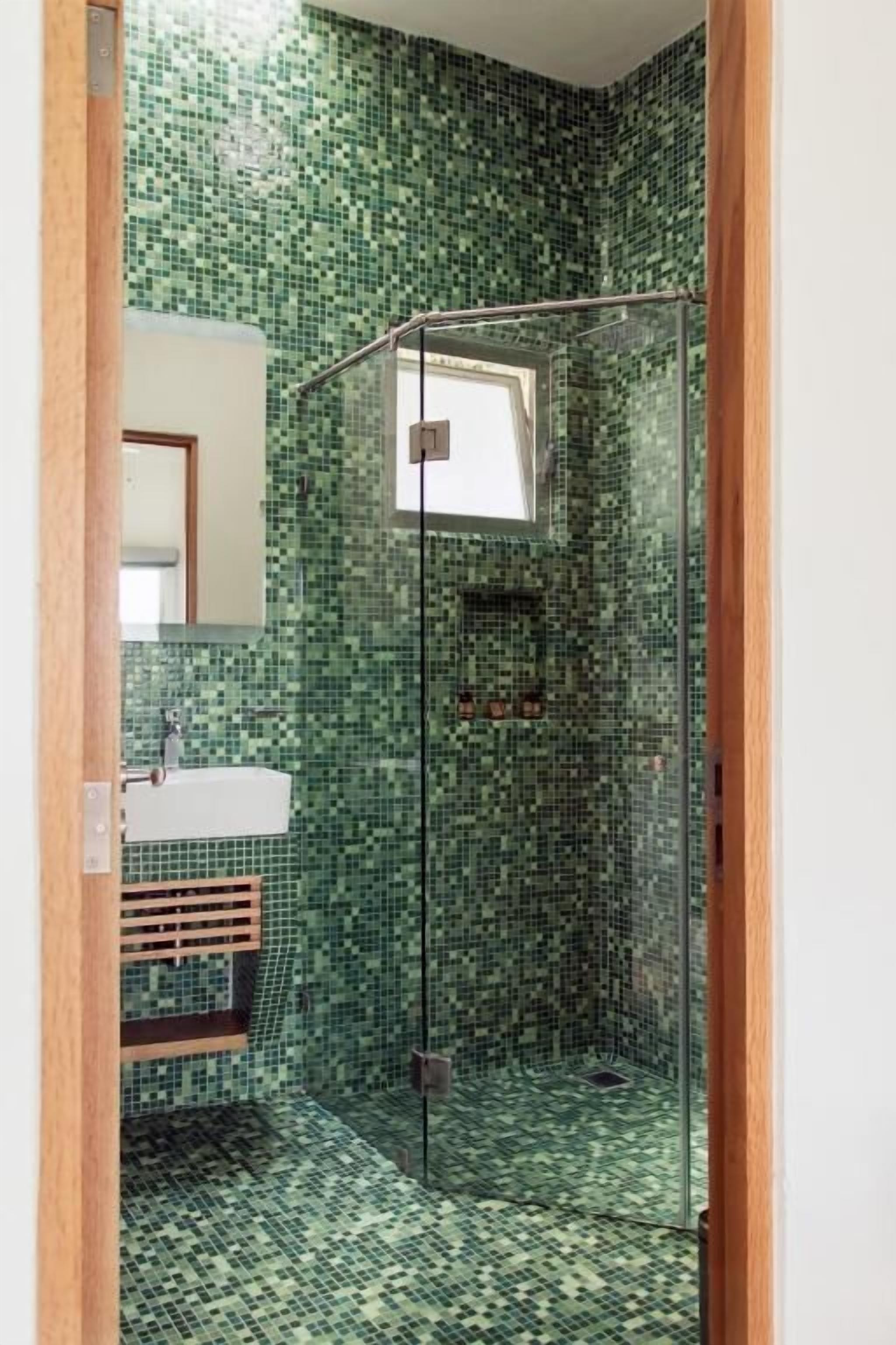 Un baño con baldosas verdes de mosaico, una cabina de ducha de vidrio y un lavamanos blanco.