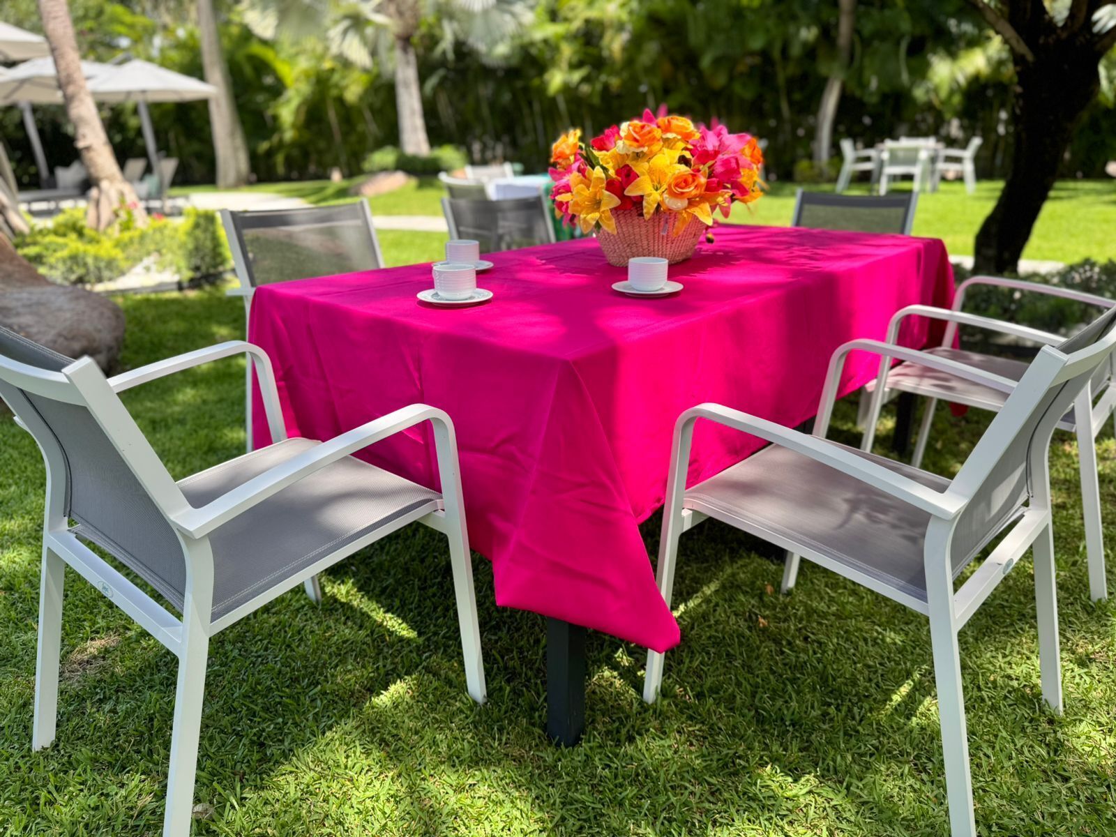 Una mesa al aire libre con mantel rosa brillante, sillas blancas y un centro de mesa floral.