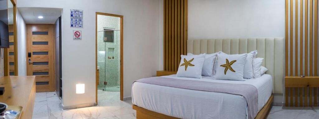 Une chambre d’hôtel avec un lit, une porte en bois, une porte en verre donnant sur une salle de bain, et une petite table avec une chaise.