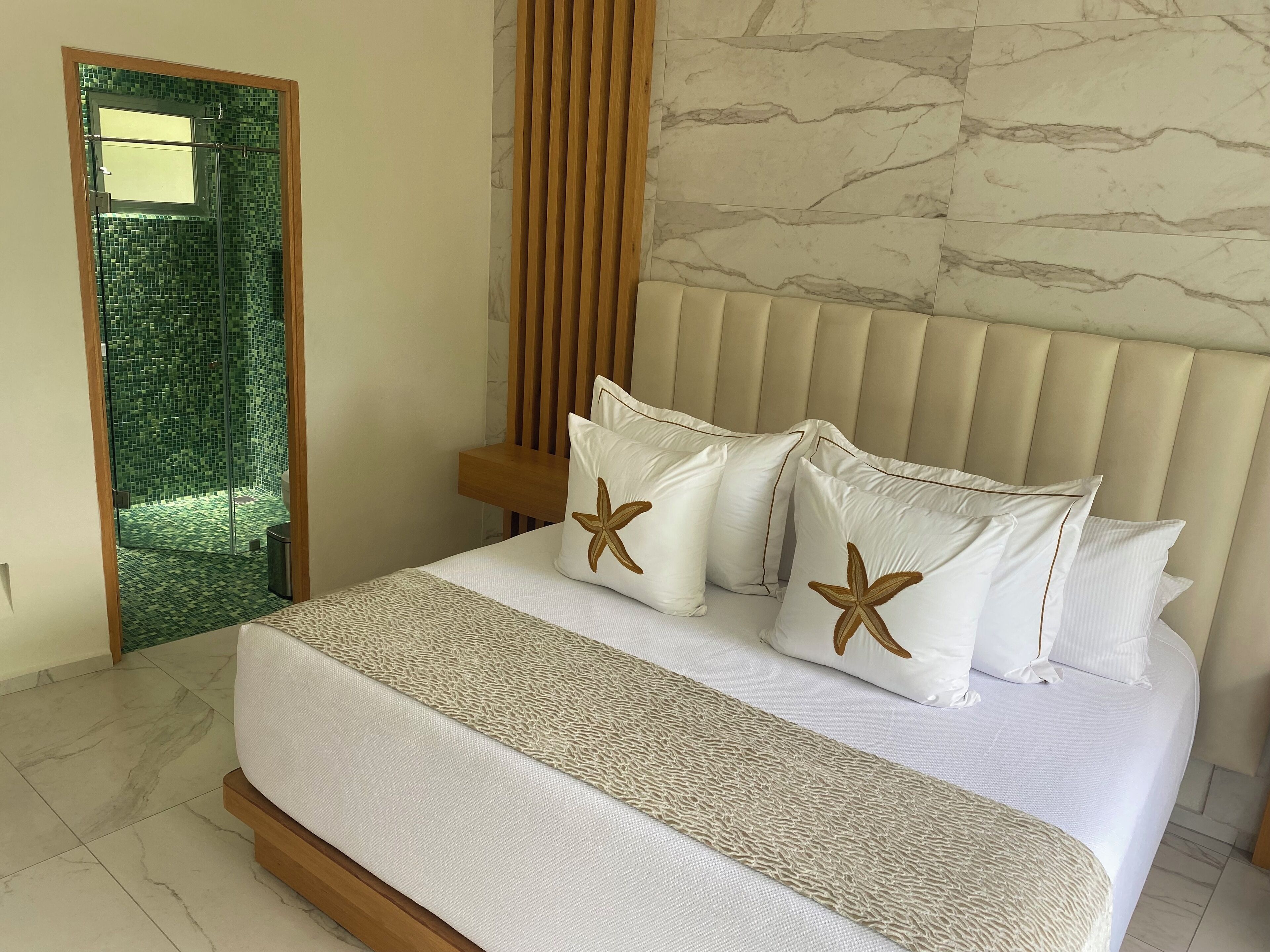 Habitación de hotel con cama, cabecera de madera, cabina de ducha con azulejos verdes y pared con diseño de mármol.