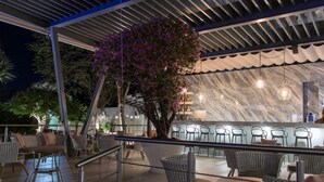 Un área exterior con mesas y sillas, un bar con asientos, y un árbol grande con flores rosadas.