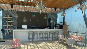 Un moderno bar con techo de madera, luces colgantes en forma de estrella y una pantalla grande que muestra música. Hay taburetes de bar y asientos acolchados con cojines estampados.