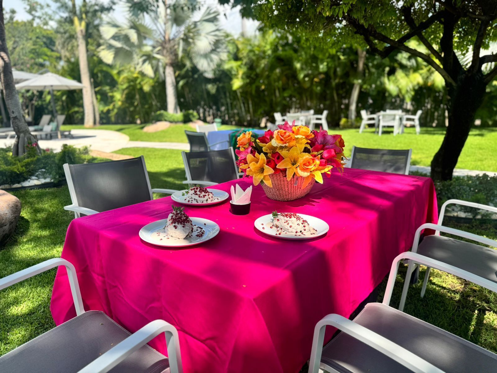 An outdoor dining area with a table covered in a bright pink tablecloth, surrounded by chairs. There are plates with food and a basket of flowers on the table.