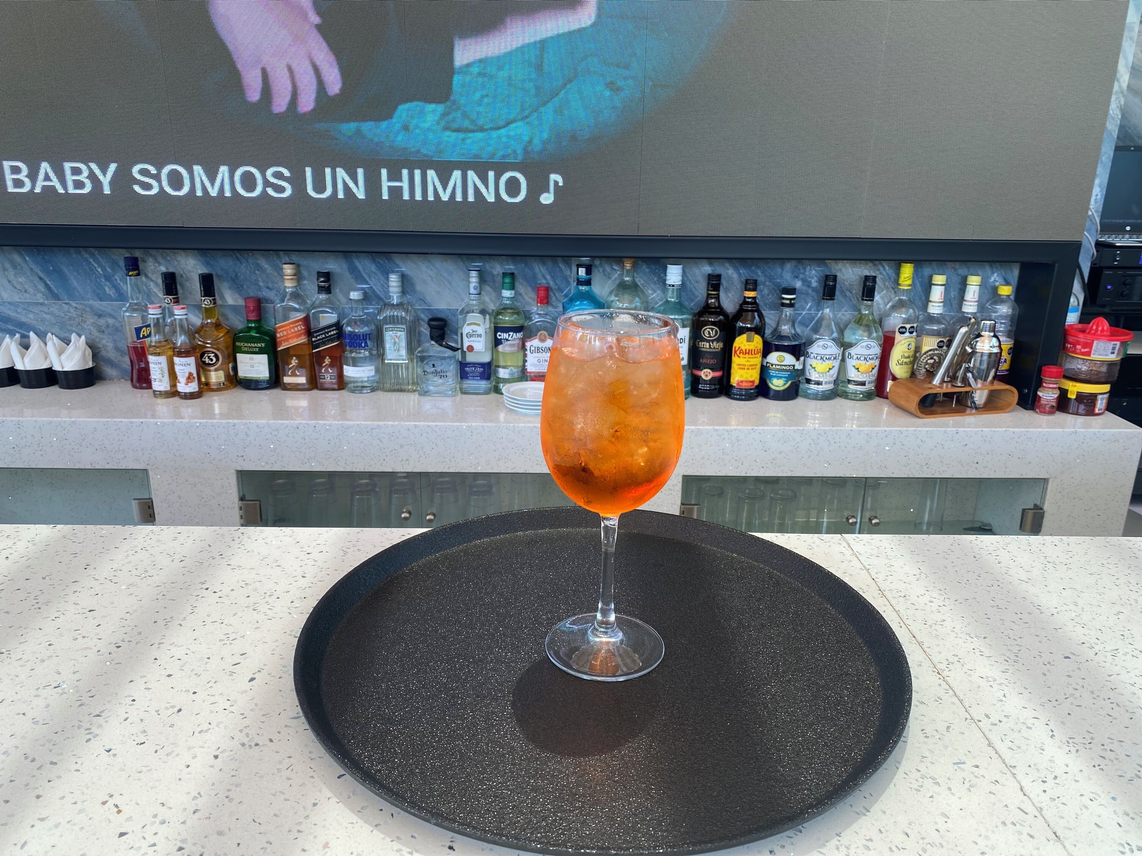 Uma área de bar com diversas garrafas de bebidas e um copo de drink vermelho sobre um apoio de bar preto.