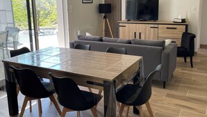 A modern living room with a wooden dining table, black chairs, a grey sofa, and a TV on a wooden cabinet. - Gîte la Chotinière (Mareuil-sur-Cher)