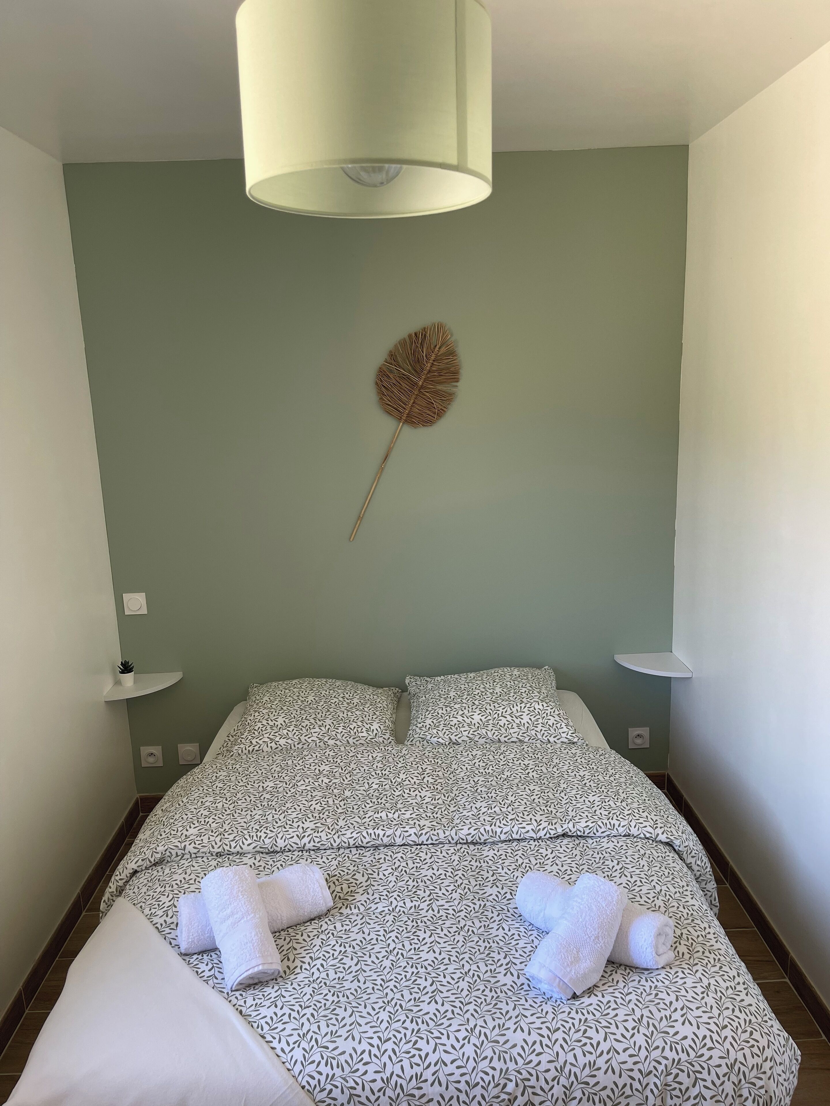 Una cama bien hecha con ropa de cama estampada y dos toallas enrolladas, una lámpara fijada a la pared y una flor seca decorativa en la pared.