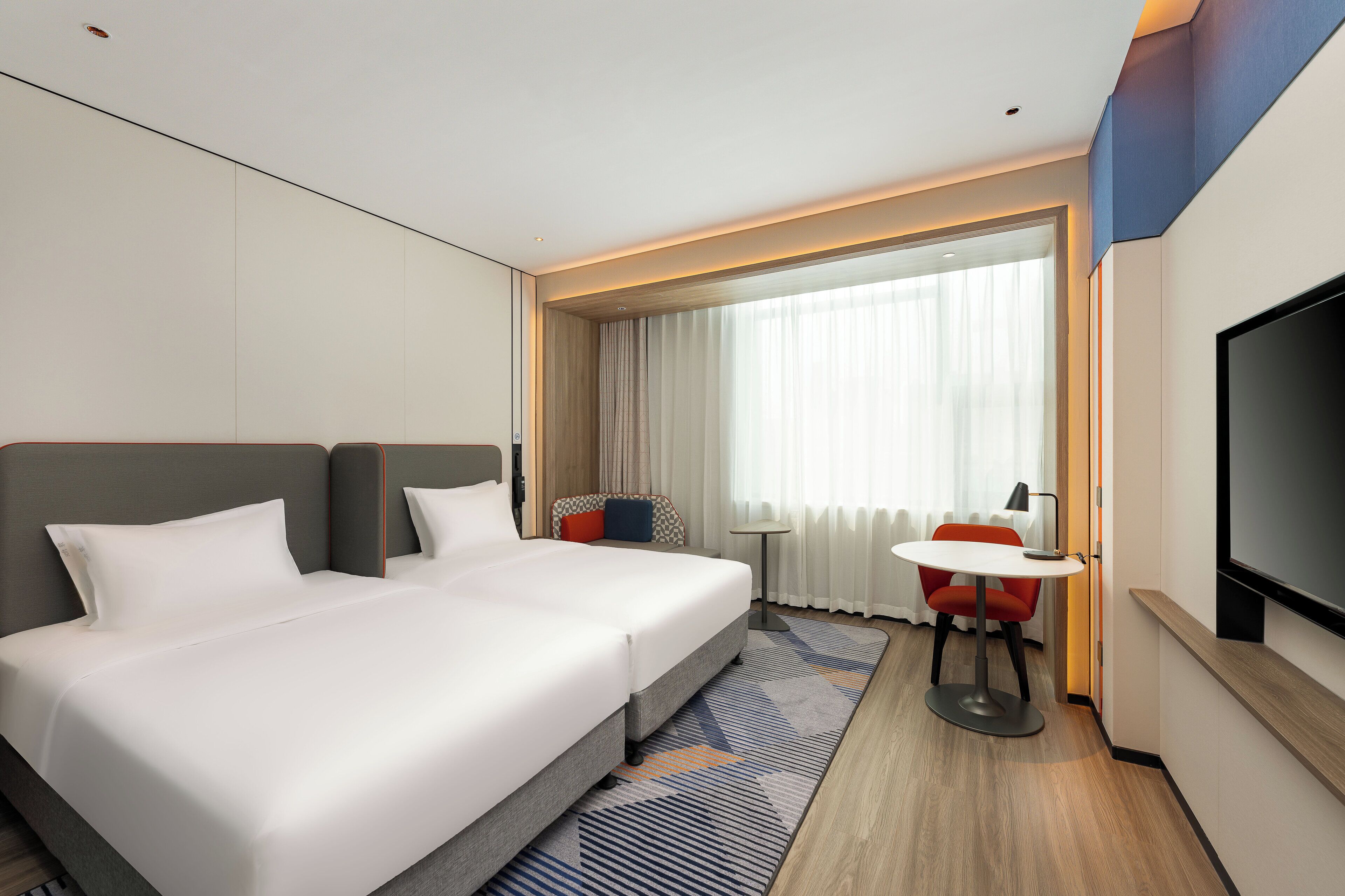 Ein Hotelzimmer mit einem großen Bett, einem kleinen Tisch, einem Fernseher und einem Fenster mit Vorhängen.