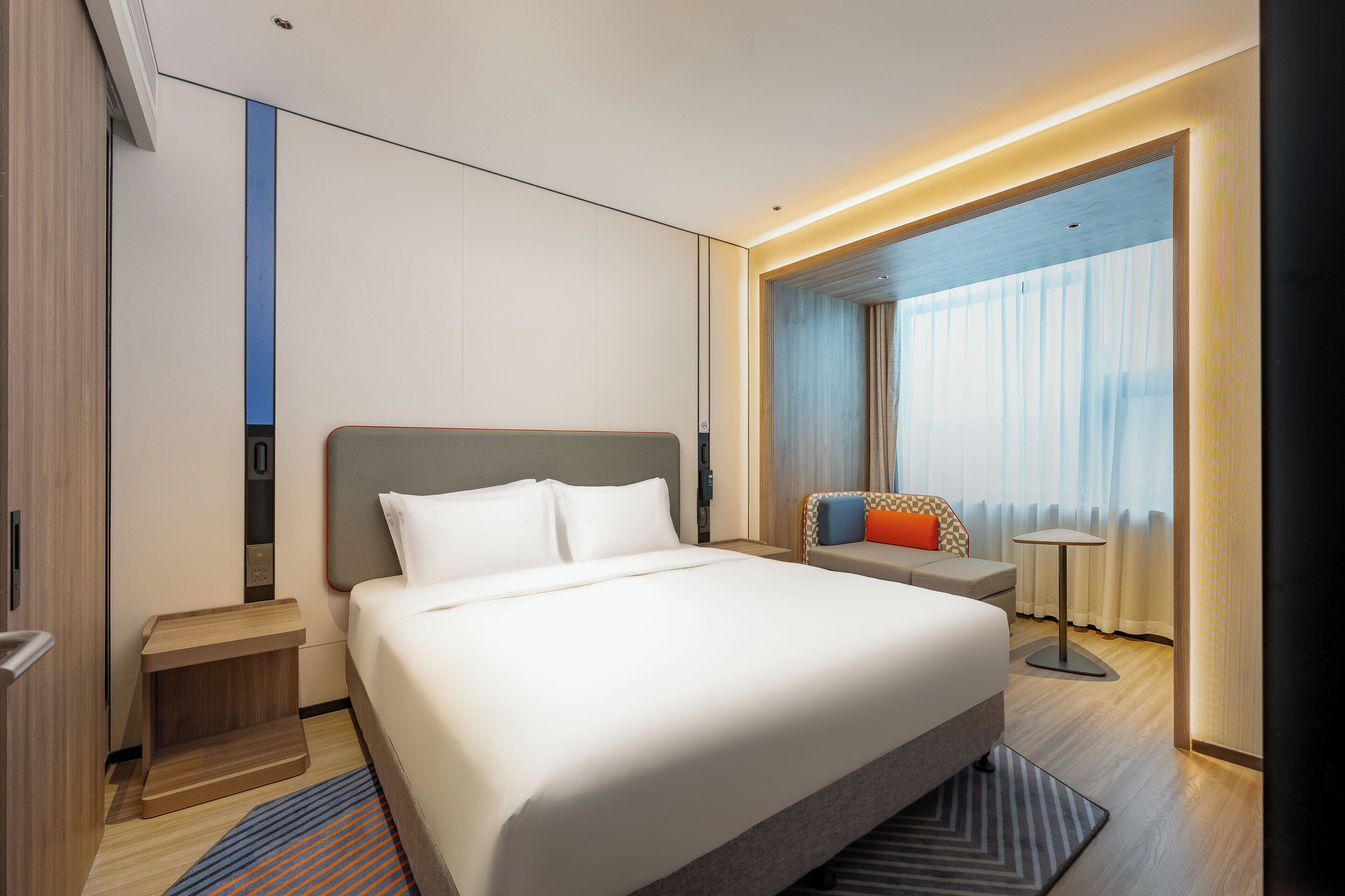 A modern hotel room with a large bed, a bedside table, a small seating area, and a view of the outdoors through a window.
