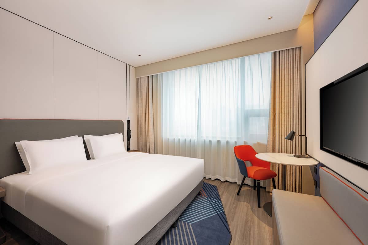 A hotel room with a large bed, a TV, a desk with a chair, and a window with curtains.