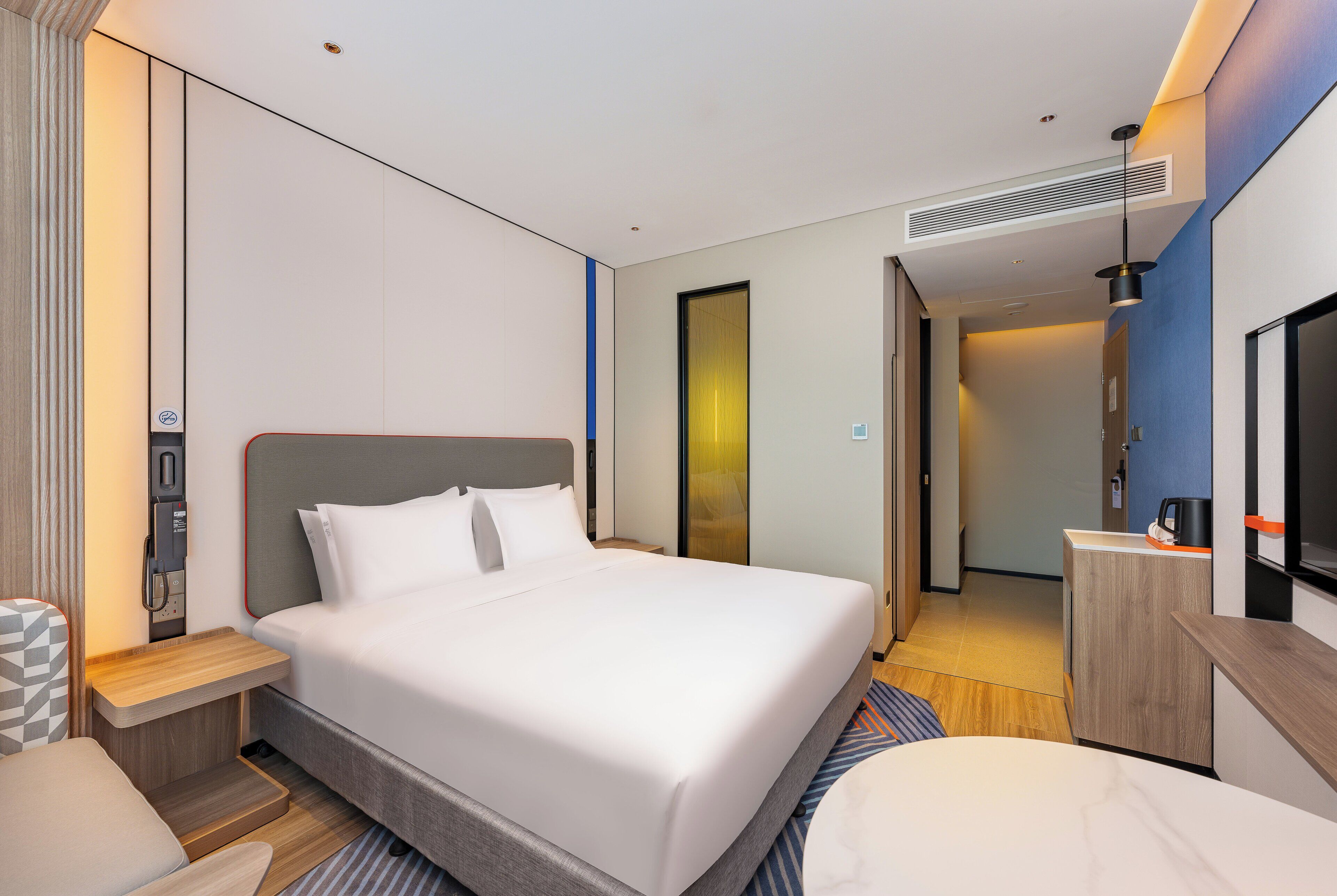 A modern hotel room with a large bed, bedside table, and a small desk area.