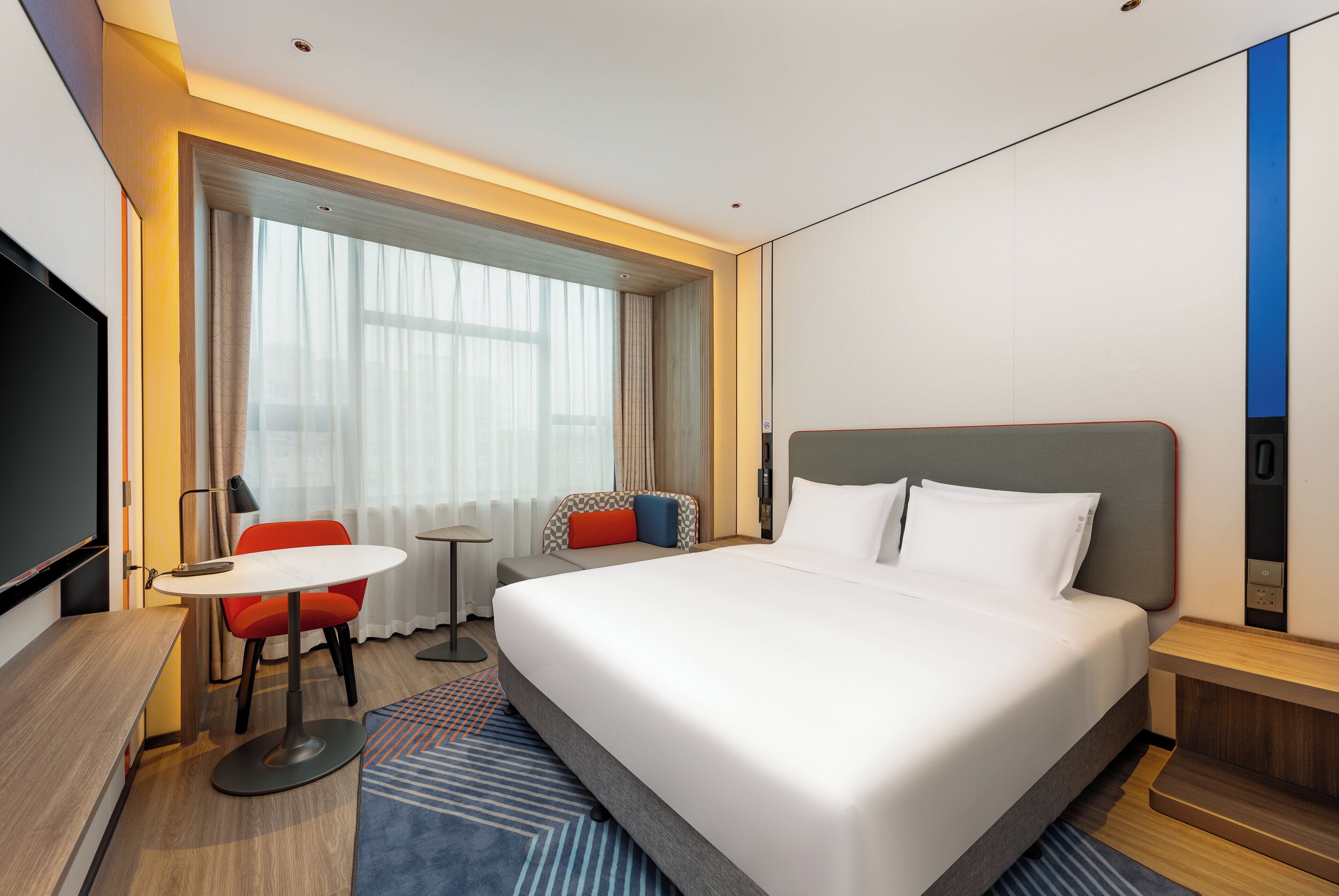 Ein Hotelzimmer mit einem großen Bett, einem Schreibtisch mit Stuhl, einem Fernseher und einem Fenster mit Vorhängen.