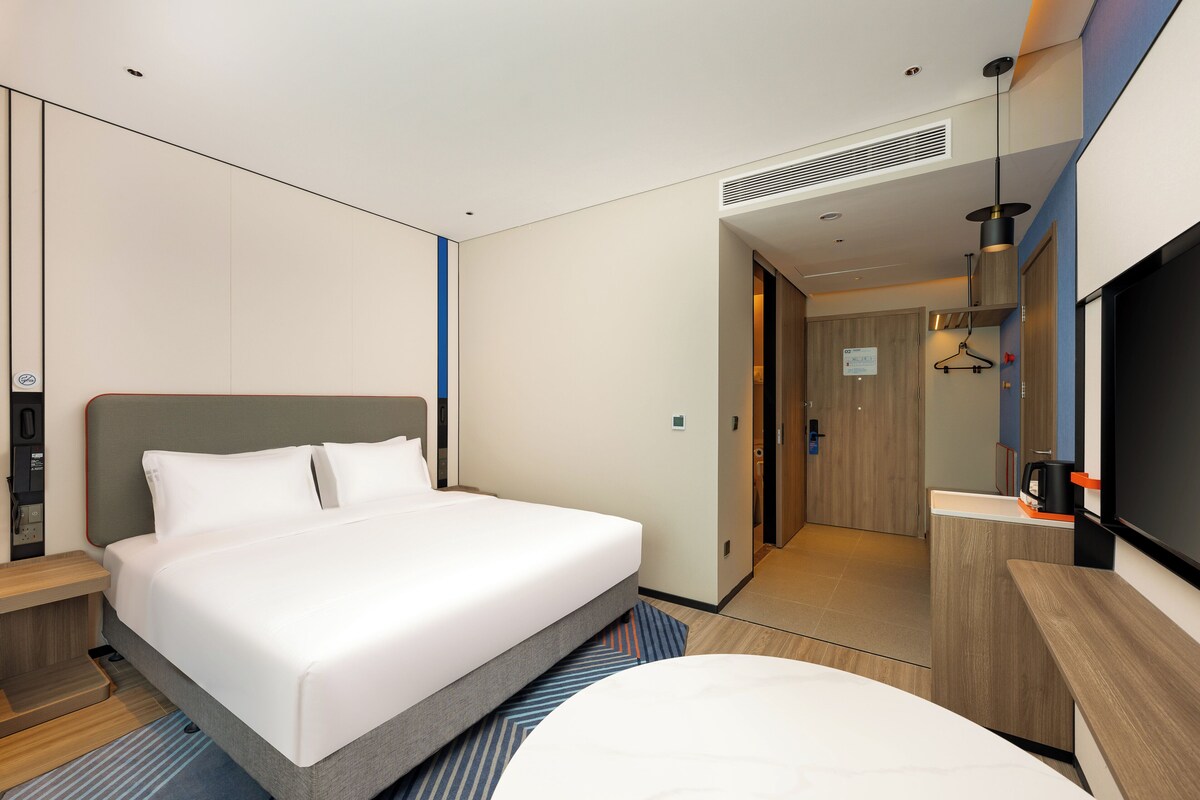 A modern hotel room with a large bed, a desk, a TV, and a bathroom visible through an open door.