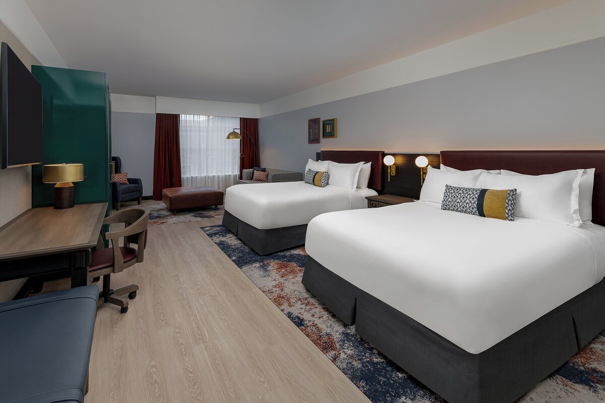 A hotel room with two beds, a desk, a chair, a lamp, and a TV.