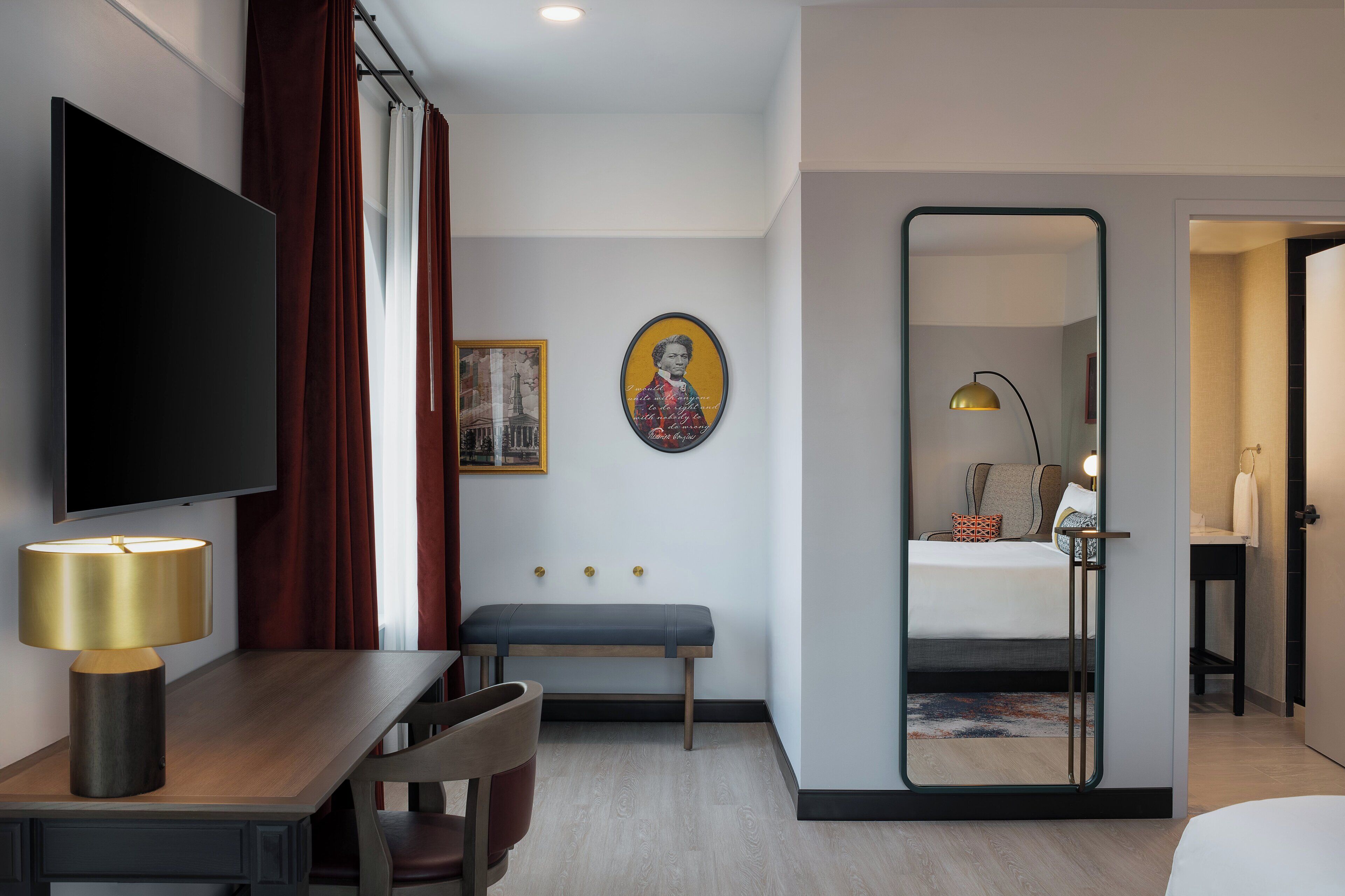 A hotel room with a flat-screen TV, a desk with a lamp, a bench, a bed, a mirror, and a painting on the wall.