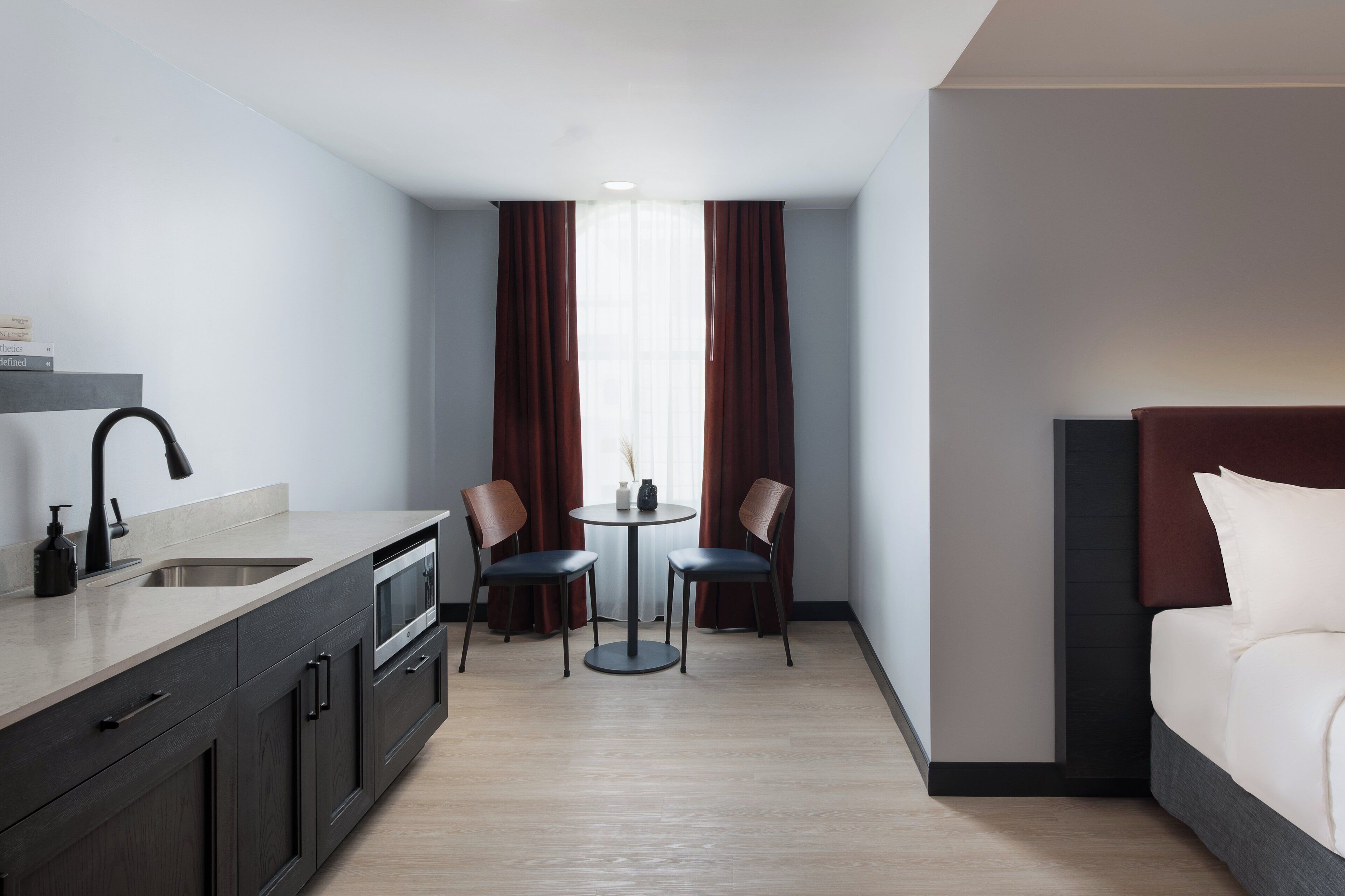 A hotel room with a bed, a kitchenette, a dining area with two chairs and a table, and a window with curtains.