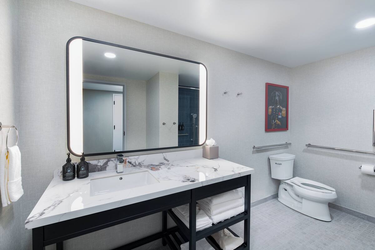 Un baño moderno con un espejo grande, lavamanos de mármol blanco, tocador negro, inodoro blanco y un perchero.
