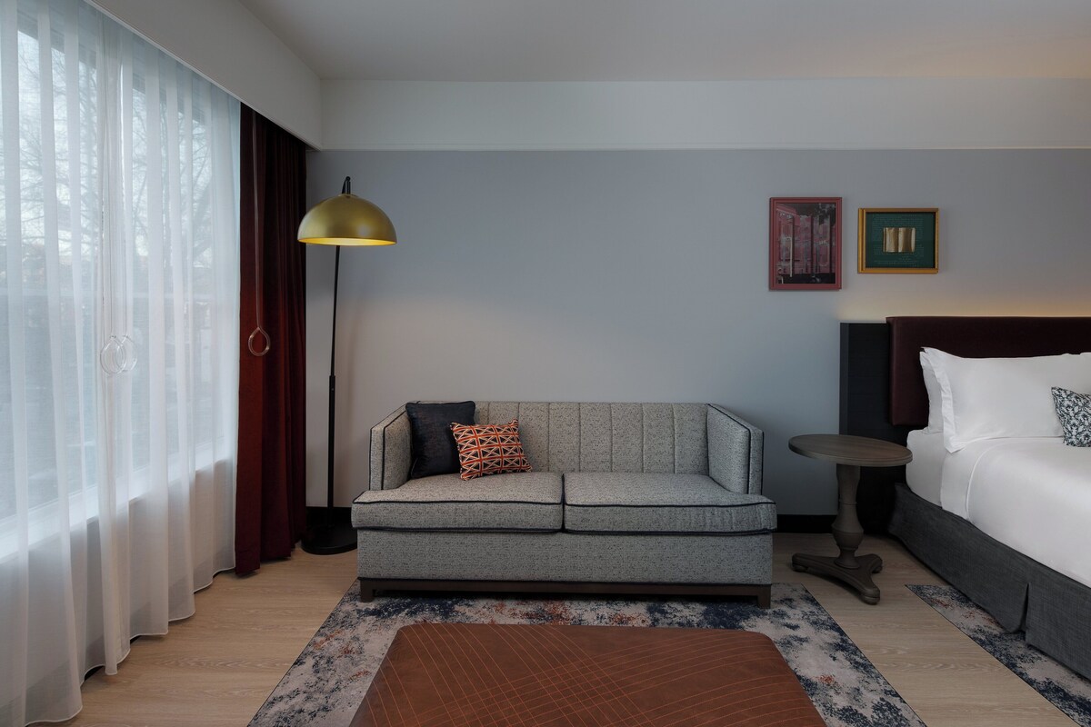 A hotel room with a grey sofa, a round side table, a bed with white bedding, and a large window with sheer curtains.