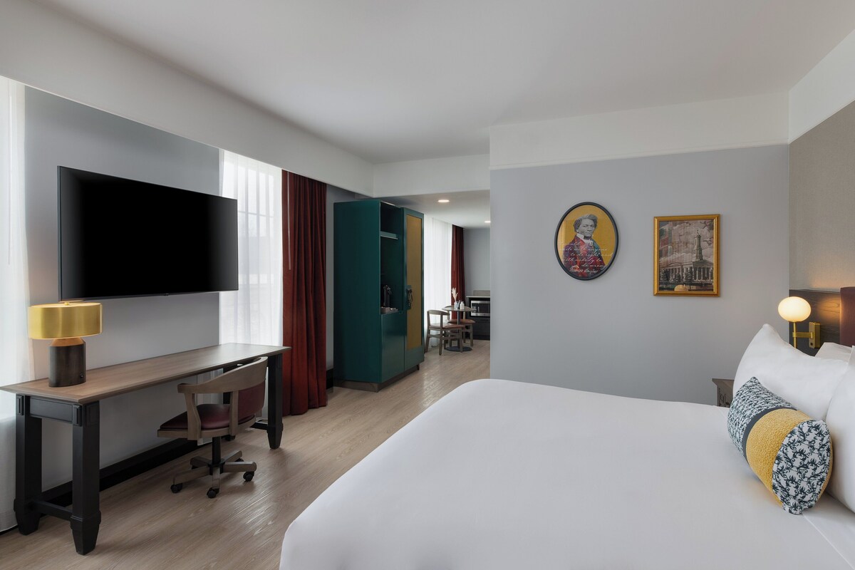 A hotel room with a bed, a desk with a chair, a television, and a dining area in the background.