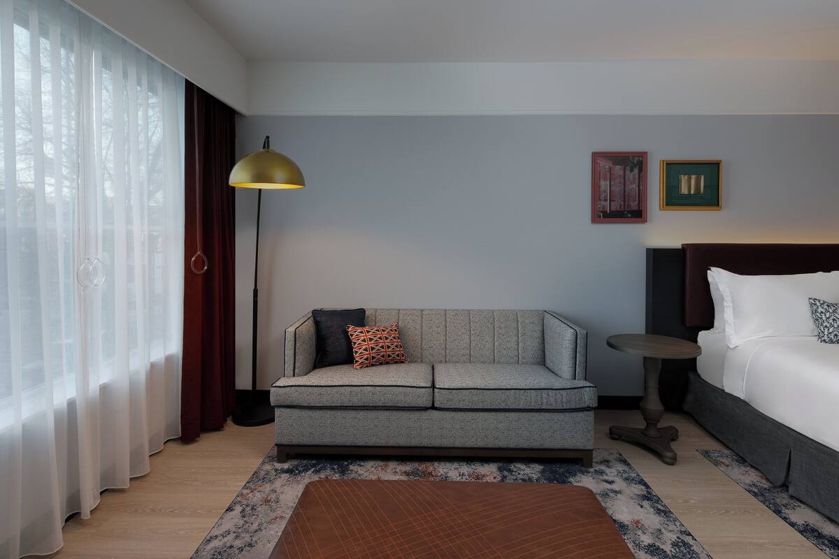 Habitación de hotel con un sofá gris, una mesita redonda, una cama con ropa blanca y un ventanal con cortinas translúcidas.