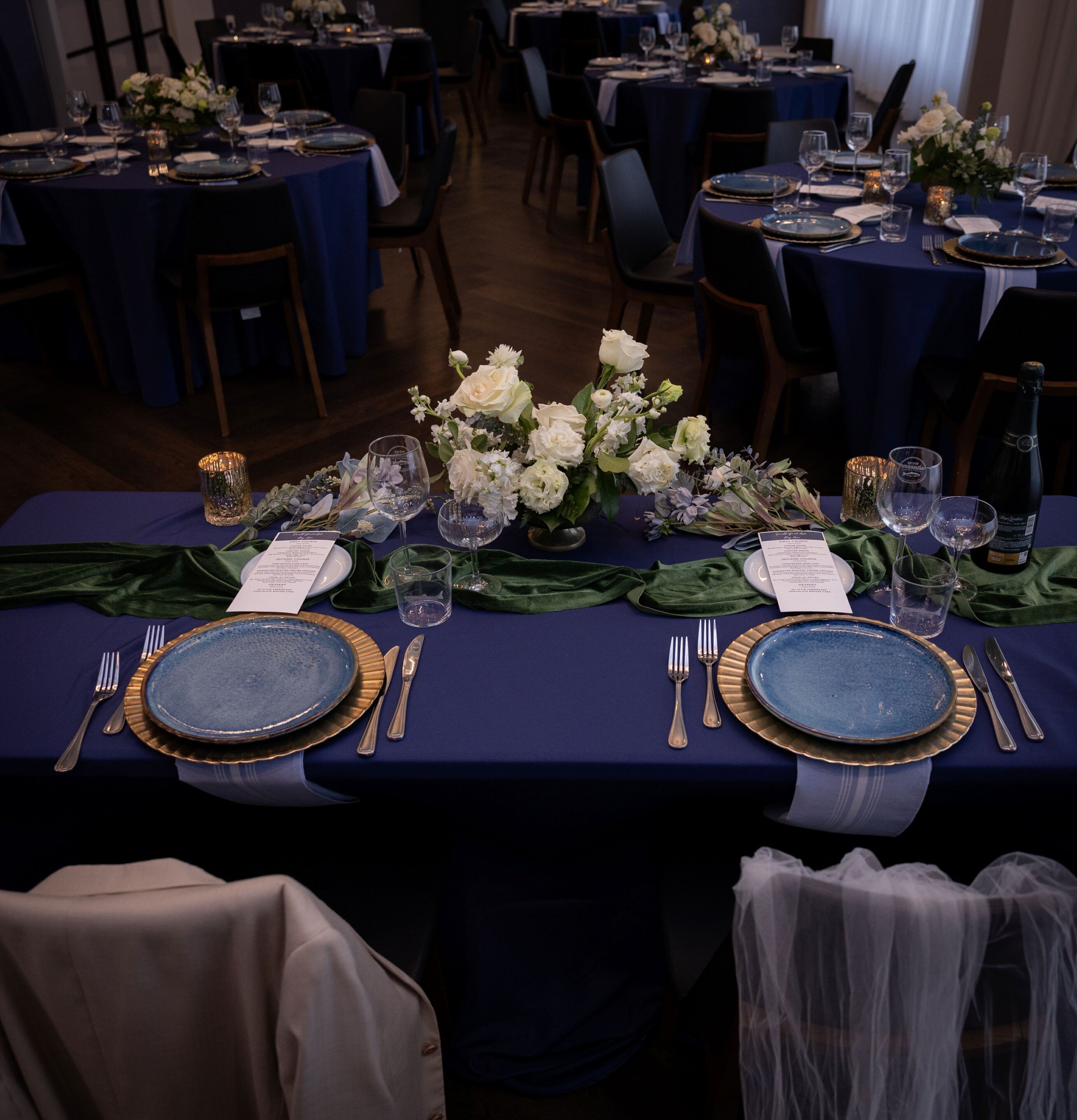 Un salón de banquetes con mesas dispuestas para un evento formal, con manteles azules, platos con detalles dorados y centros de mesa florales.
