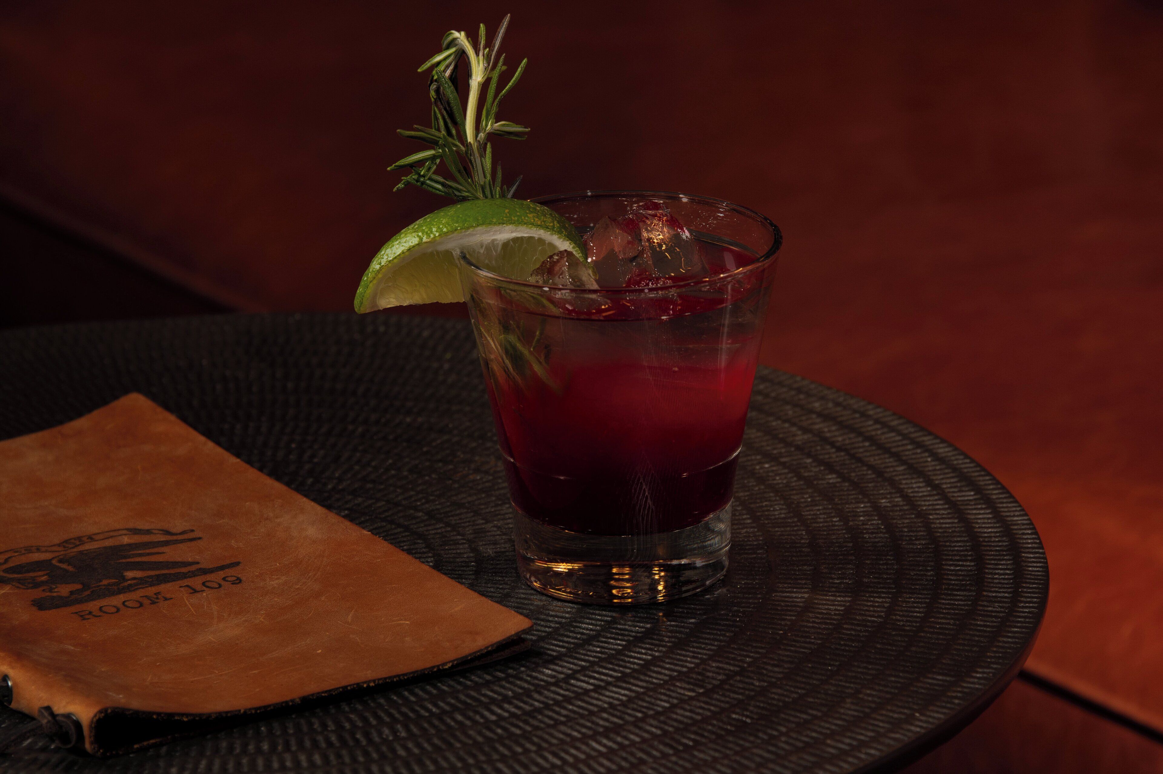 A glass of red drink with a lime wedge and a sprig of rosemary on a coaster.