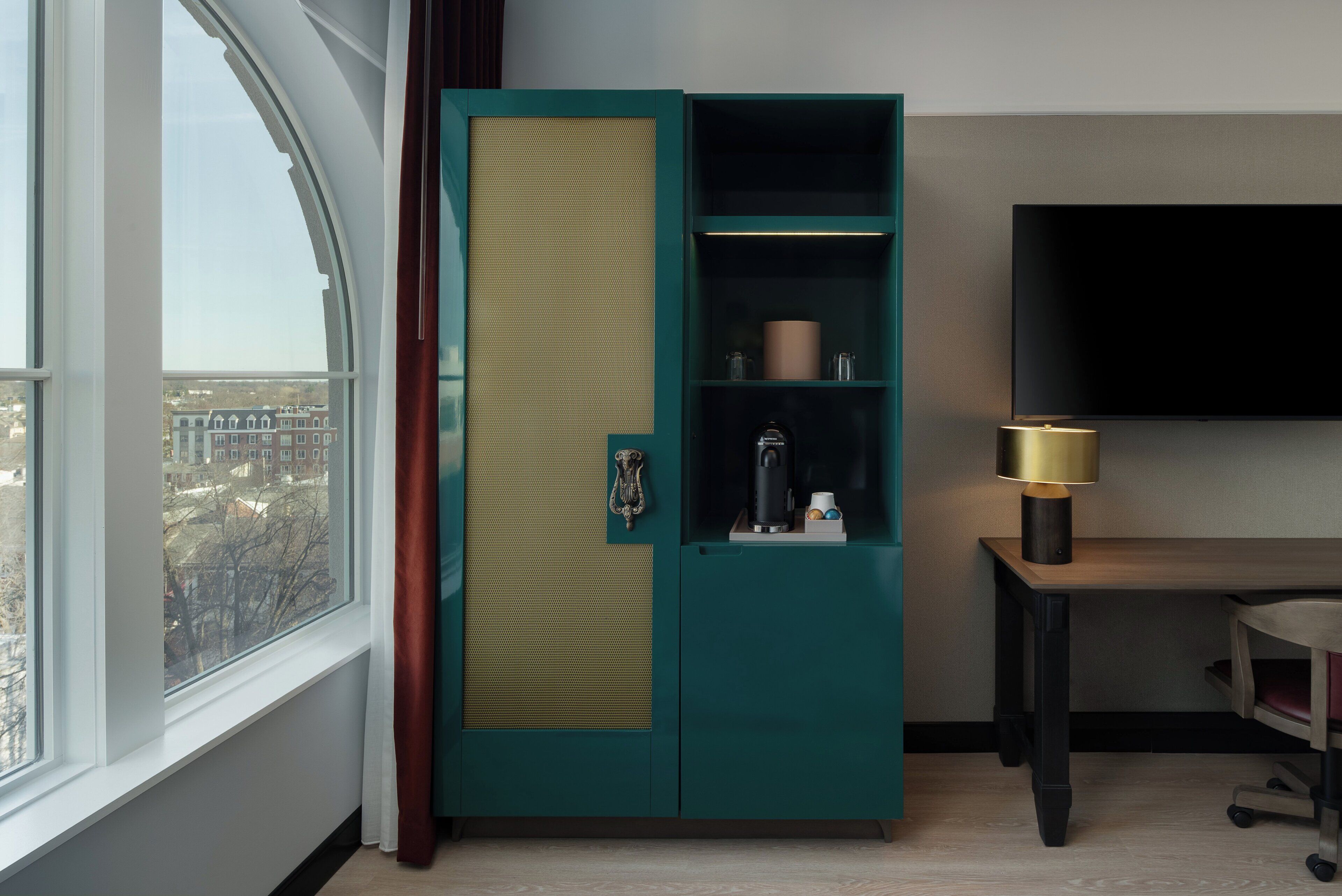 A teal wardrobe with a mirrored section, a television mounted on the wall, and a desk with a lamp.
