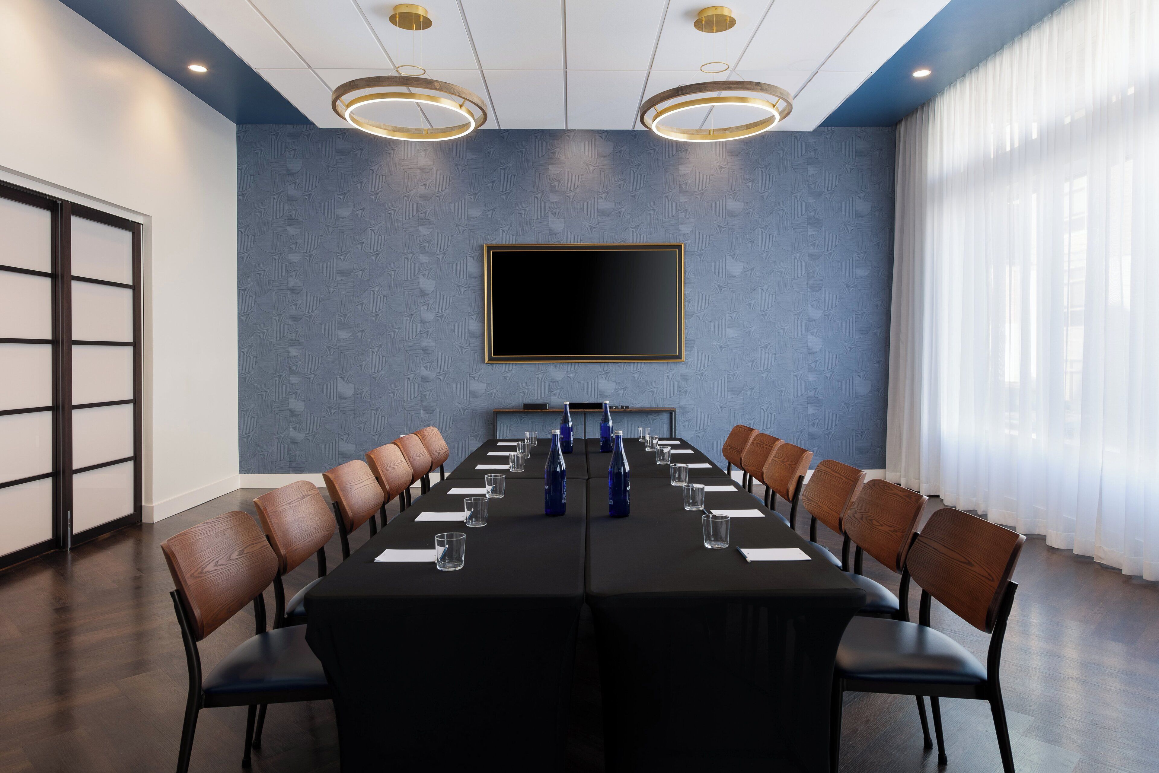 Une salle de conférence avec une longue table, des chaises, un grand écran et une fenêtre ornée de rideaux légers.